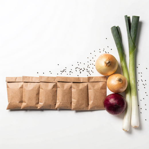 Brown paper seed packets placed beside yellow and red onions and two young leeks